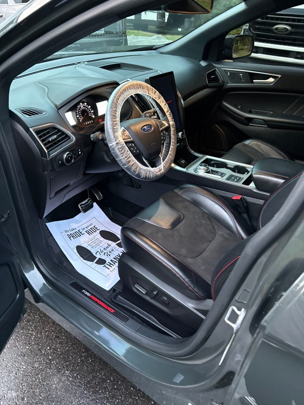 Interior of Ford sedan showing steering wheel with protective cover, black leather seats, dashboard, and driver-side door open with window sticker visible
